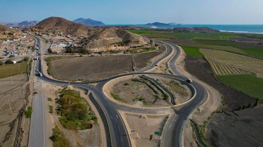 La imagen tomada por la SNCI muestra una rotonda en una vía de conexión con un paisaje costero al fondo. Se observa una infraestructura vial en desarrollo o recientemente construida, con áreas aledañas de cultivo y una zona urbana en crecimiento.