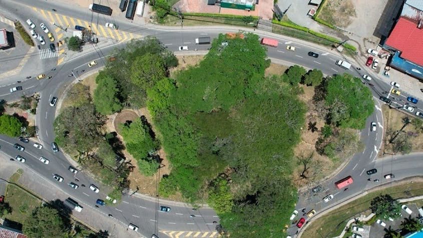 La imagen muestra una vista aérea de una gran rotonda con abundante vegetación en el centro, rodeada por múltiples carriles de tráfico. Se observa la circulación de vehículos en diferentes direcciones, con señalización vial visible en las intersecciones.