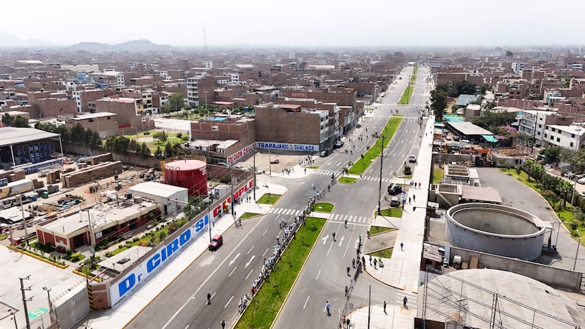 Vista aérea de la avenida Carlos Izaguirre en el Callao, Perú, mostrando una vía moderna con separador central con áreas verdes, señalización peatonal y edificios residenciales y comerciales a ambos lados. Se observan peatones y vehículos transitando en un entorno urbano denso.
