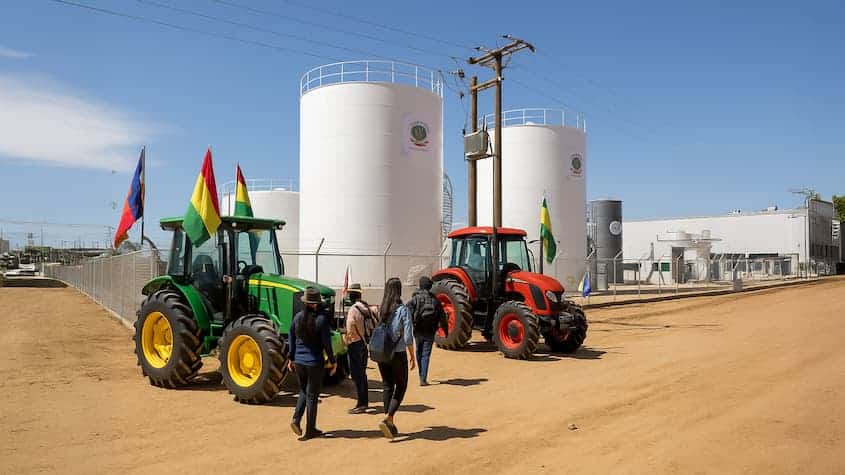 Vista de una planta de biodiésel en Bolivia, con tanques de almacenamiento blancos, tractores con banderas nacionales y visitantes caminando por un camino de tierra. Al fondo se observan instalaciones industriales modernas, cercadas y señalizadas, bajo un cielo despejado.