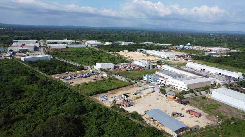 Vista aérea de un parque industrial en Colombia, rodeado de vegetación y con múltiples naves logísticas, almacenes y zonas de carga. Se observan camiones, áreas de almacenamiento al aire libre y estructuras metálicas distribuidas en un entorno amplio y ordenado, bajo cielo parcialmente nublado.