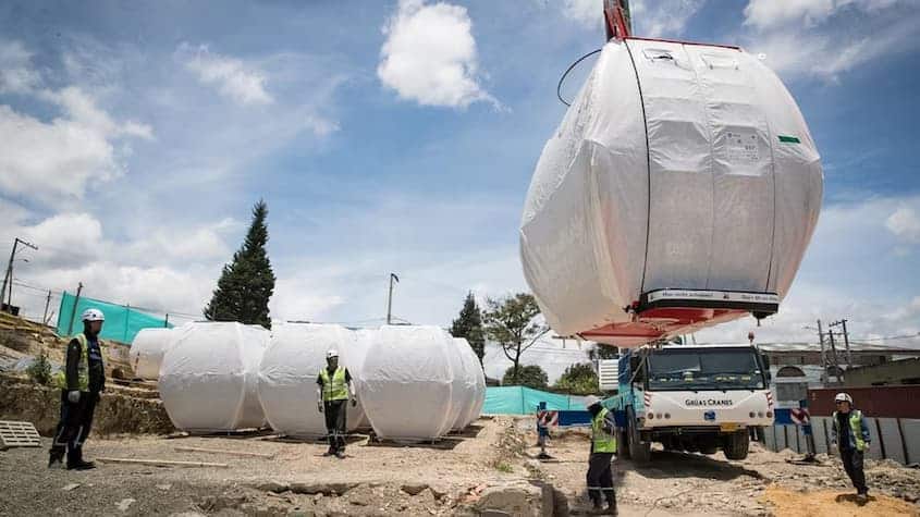 Personal técnico supervisa la instalación de cabinas envueltas del sistema de transporte TransMiCable en San Cristóbal, Bogotá. Una grúa descarga una cabina desde un camión, mientras otras esperan en el suelo. La escena ocurre en un terreno en preparación, bajo cielo parcialmente nublado.
