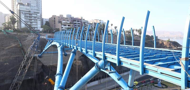 Vista del puente peatonal metálico en construcción como parte del corredor turístico Miraflores–Barranco en Lima, Perú. La estructura de color azul destaca por su diseño moderno sobre los acantilados de la Costa Verde, conectando dos distritos urbanos con vista al mar y rodeado de edificios residenciales.