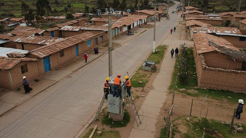 Técnicos instalan un transformador eléctrico en un poste dentro de una comunidad rural del Cusco como parte de un proyecto de electrificación trifásica. Las viviendas de adobe con techos de tejas rojas y los caminos asfaltados evidencian el entorno andino. Se observa a pobladores transitando por la zona mientras se realiza la obra.