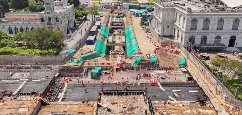 Vista aérea de las obras de construcción de la Estación Central de la Línea 2 del Metro de Lima, ubicadas entre históricos edificios coloniales. Se observa maquinaria, estructuras de concreto, obreros trabajando y lonas verdes cubriendo sectores de la excavación, en pleno centro de la ciudad.