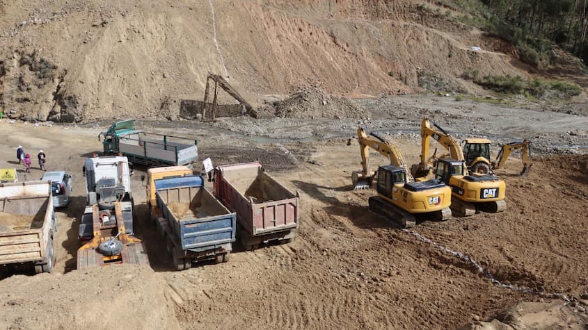 Obras de construcción de la represa Vacas Kuchu-Chilawaki, con maquinaria pesada en operación, incluyendo excavadoras CAT y varios volquetes de carga en una zona de movimiento de tierras. El entorno montañoso y el terreno agreste evidencian el trabajo de infraestructura hidráulica en una región rural.