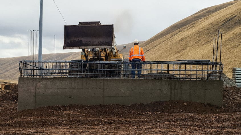 Obrero supervisa construcción de base para puente modular en zona rural andina, con maquinaria pesada y estructura metálica en avance.